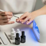 Osoba wykonująca manicure hybrydowy: aplikacja lakieru pędzelkiem, narzędzia oraz włączona lampa UV na stanowisku pracy.