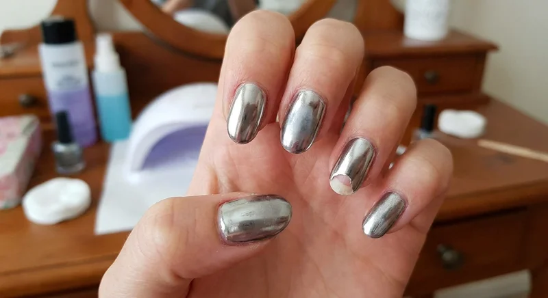 Dłoń ze srebrnym, lustrzanym manicure, z widocznym dużym odpryskiem pyłku u nasady jednego z paznokci.