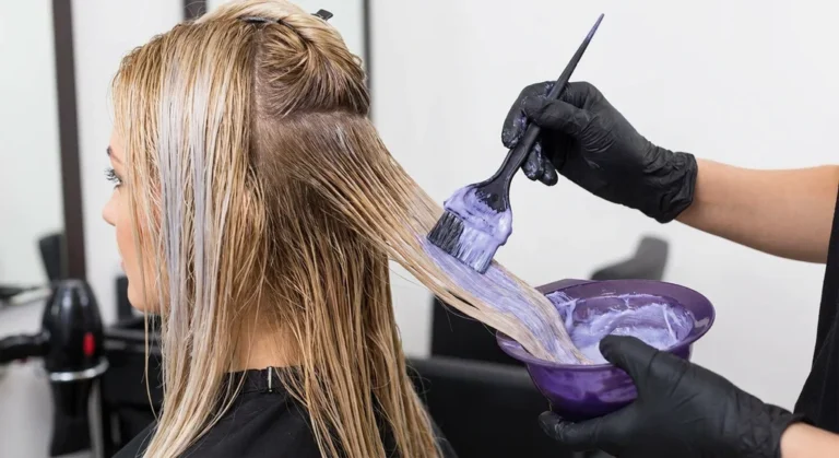 Fryzjer nakłada pędzelkiem fioletowy toner na blond włosy w celu neutralizacji żółtych tonów.